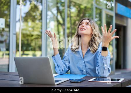 Enttäuschte junge Geschäftsfrau, die sich frustriert fühlte, mit einem Laptop draußen auf der Straße saß, aufschaute und sich beschwerte Stockfoto