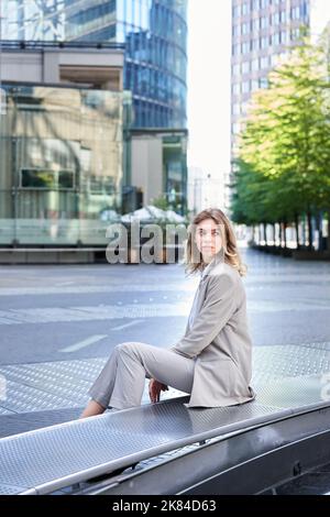 Weibliche Angestellte, die im Freien in der Nähe von Bürogebäuden sitzt, dreht sich um und schaut nach hinten und wartet auf die Mitarbeiterin in der Mittagspause Stockfoto