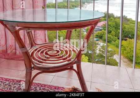 Innenraum des Balkons mit Korbtisch mit Glasdeckel, Blick vom Balkon auf die Küste, rosa getönte Farben. Stockfoto