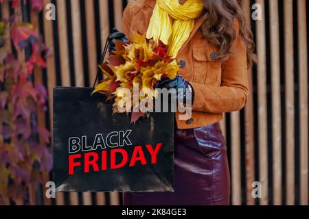 Hallo november. Nahaufnahme einer trendigen Frau mittleren Alters in orangefarbenem Trenchcoat mit Einkaufstasche und herbstgelben Blättern in der Stadt. Stockfoto
