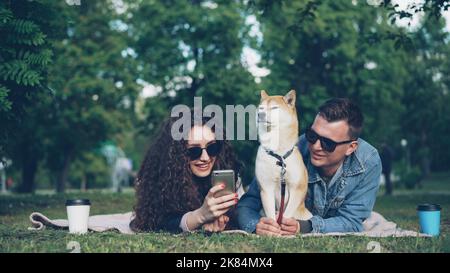 Die junge Frau zeigt ihrem fröhlichen Freund lustige Bilder auf dem Smartphone, während sie sich im Park mit einem Hund entspannt, die Menschen liegen auf karierten Wiesen und beobachten den Bildschirm. Stockfoto