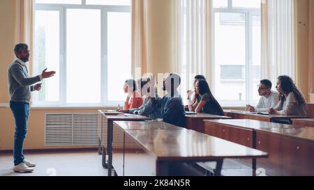 Ein freundlicher Lehrer spricht mit Schülern, ein kluger Mann hebt die Hand und spricht am Tisch in einem geräumigen Hörsaal, während seine Klassenkameraden schreiben. Stockfoto