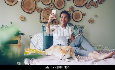 Ein glücklicher afroamerikanischer Teenager telefoniert mit einem Smartphone, das Freunde anruft, die reden und gestikulieren, während ihr niedlicher Hund mit ihr auf dem Bett liegt. Stockfoto