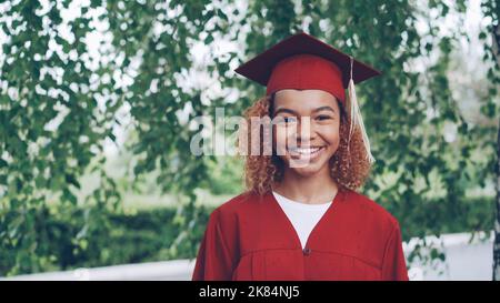 Porträt eines hübschen afroamerikanischen Mädchens glücklich Abschluss Student in rotem Kleid und Mortarboard im Freien stehend, lächelnd und Blick auf die Kamera. Jugend- und Bildungskonzept. Stockfoto