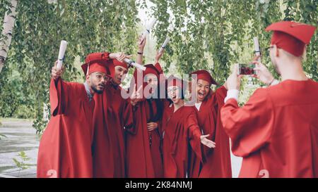Der graduierende Student nimmt ein Video seiner Freunde in Kleidern und Leichenbrettern auf, die Diplome halten, die Hände winken und posieren und auf die Smartphone-Kamera blicken. Menschen- und Technologiekonzept. Stockfoto