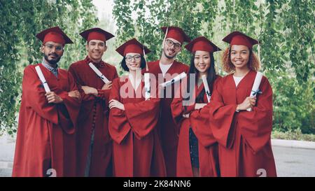 Portrait einer multirassischen Gruppe von Absolventen, die Diplome halten und mit gekreuzten Armen im Freien stehen, Kleider und Mörsertafeln tragen, lächeln und die Kamera betrachten. Stockfoto