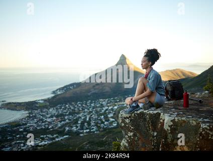 Es ist eine ziemliche Steigung, die versucht, Ihre Fitness-Ziele zu erreichen. Eine Frau, die auf die Aussicht schaut, während sie auf einer Bergklippe sitzt. Stockfoto