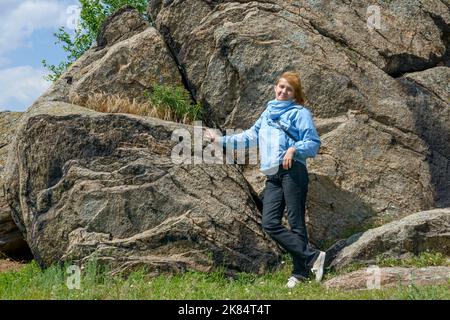 Ältere Frau in blauer Jacke posiert in der Nähe von großen abgespaltenen Felsbrocken auf dem Wildfeld am Ufer des Dnjepr-Flusses in der Ukraine. Stockfoto