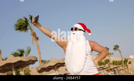 Der Weihnachtsmann macht Selfie. Weihnachtsmann Sommerurlaub. Weihnachtsmann-Blogger. Funny Santa, in Sonnenbrillen, nimmt Selfie-Video am Strand am Meer, vor Palmen Hintergrund. Hochwertige Fotos Stockfoto