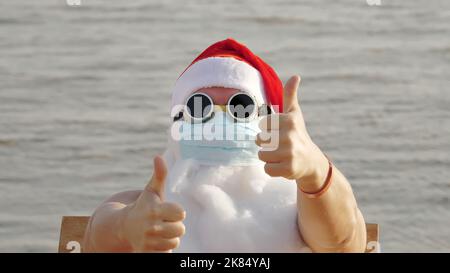 Witziger weihnachtsmann, in einer Schutzmaske. Weihnachtsmann, in Sonnenbrillen, winkende Hände, am Strand am Meer, entspannen. weihnachtsmann Urlaub während Coronavirus Pandemie oder Lockdown, an der Küste. Hochwertige Fotos Stockfoto