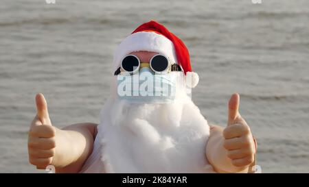 Witziger weihnachtsmann, in einer Schutzmaske. Weihnachtsmann, in Sonnenbrillen, winkende Hände, am Strand am Meer, entspannen. weihnachtsmann Urlaub während Coronavirus Pandemie oder Lockdown, an der Küste. Hochwertige Fotos Stockfoto
