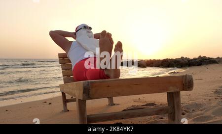 Lustige santa claus Sonnenbäder. santa, in Sonnenbrillen, liegt auf Holzliege am Strand am Meer, entspannend. santa claus ist im Sommerurlaub, an der Küste. Hochwertige Fotos Stockfoto