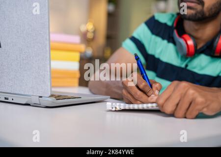 Nahaufnahme eines Mannes, der während des Online-Unterrichts oder Tutoriums zu Hause mit einem Laptop Notizen schreibt - Konzepte von E-Learning, Fernunterricht und Internet. Stockfoto