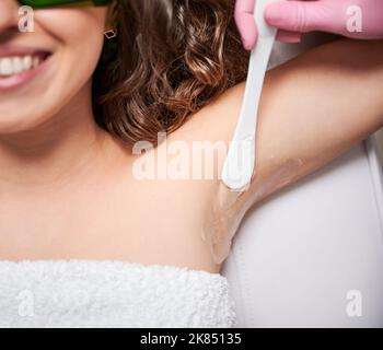 Nahaufnahme des Kosmetikers, der vor dem Laserepilieren Gel auf den weiblichen Achselbereich aufgetragen hat. Lächelnde junge Frau erhält Laser-Haarentfernung Behandlung im Schönheitssalon. Stockfoto