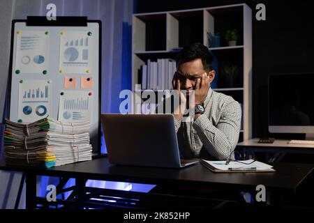 Müde oder gelangweilt asiatischen männlichen Arbeiter sitzen am Schreibtisch brauchen Ruhe leiden Müdigkeit überarbeitet im Büro. Erschöpfter Mann, der spät in der Nacht am Computer arbeitet, versucht es Stockfoto