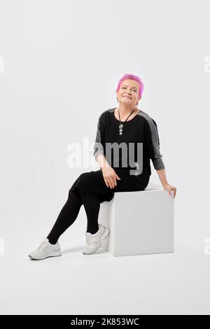 Glückliche alte Frau mit kurzen rosafarbenen Haaren in langem Strickkleid sitzt auf weißer Schachtel im Studio Stockfoto