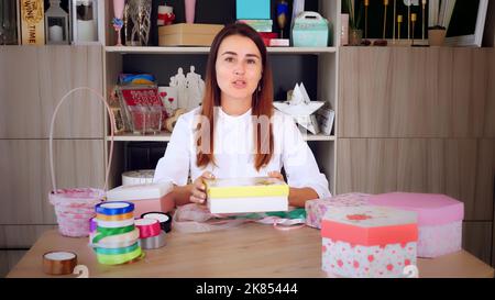 Geschenkverpackung. Attraktive Frau, Designer von Geschenken und Dekorationen zeigt Training online, zeichnet Videoblog, Webinar, sitzt vor dem Smartphone, Kamera. Online-Verkauf. Hochwertige Fotos Stockfoto