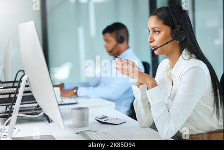 Sehen Sie die Seite, über die im spricht? Eine junge Callcenter-Agentin, die an ihrem Schreibtisch sitzt und einem Kunden erklärt, während sie ein Headset trägt. Stockfoto