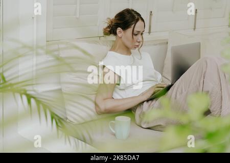 Junge Frau, die zu Hause auf der Terrasse an einem Laptop arbeitet. Die junge Frau liegt auf einer Bank auf der Terrasse zu Hause und arbeitet an ihrem Laptop. Digitale Nomade Stockfoto