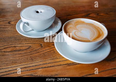 Zwei Cafe Latte mit Milchherz und Schaumkaffeebohnen in weißen Tassen auf einem Holztisch. Hintergrund der morgendlichen Energieerhöhung Stockfoto