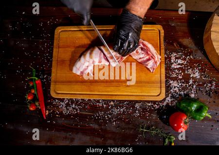 Gesalzener Schmalz, rohes Schweinefleisch mit Gewürzen auf Holzschneidebrard Studiofoto. Ein Metzger in schwarzen Handschuhen schneidet Schmalz dünn Stockfoto