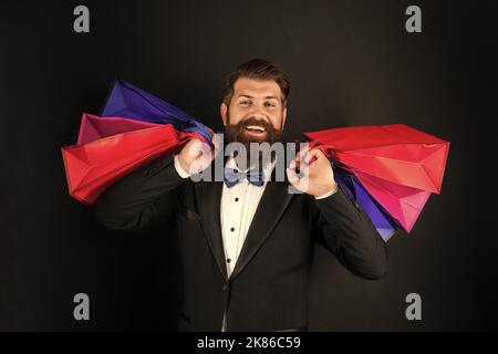 Glücklicher Mann in Smoking Fliege mit Business-Belohnung in Einkaufstaschen. Anlass Gruß. Stockfoto