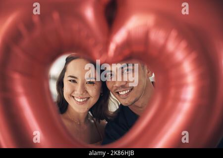 Das perfekte Bild der Liebe. Ein junges Paar posiert zu Hause durch einen herzförmigen Ballon. Stockfoto