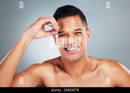 Porträt eines Mannes, der mit einer Pinzette seine Augenbraue pinkiert, im Studio mit grauem Hintergrund. Glücklich, Lächeln und junge Kerl Zupfen Stirn für Haarentfernung Stockfoto