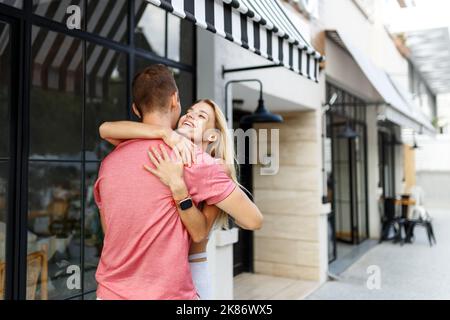 Abschiedskonzept. Erfreut glücklich lächelnde blonde Frau sagt Auf Wiedersehen oder hallo zu Freund, gibt warme Umarmung, Pose zusammen gegen Café-Platz. Wahrheitsgemäße Gefühle Stockfoto