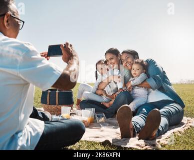 Familie, Telefon und Picknick-Foto mit Lächeln, Umarmung und glücklich zusammen im Sommer auf Gras im Park. Großeltern, Mutter oder Kinder mit Smartphone-Bild Stockfoto