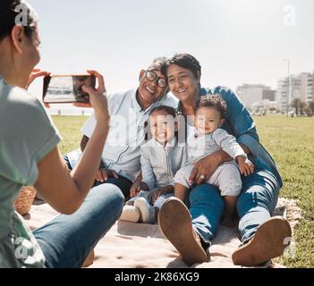 Großeltern, Kinder und Mutter mit Foto am Telefon im Sommer gemeinsam im Park. Entspannen Sie sich, glücklich und lächeln Sie von Kindern und älteren Männern und Frauen mit Stockfoto