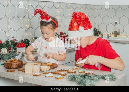 Kleine dunkelhaarige Mädchen 3 Jahre alt und Jungen 8 Jahre alt in roten Santa Hut schmücken Lebkuchen mit Glasur. Geschwister in weißen Weihnachten Stockfoto