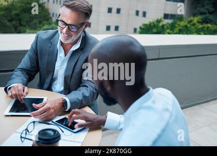 Es gibt eine App, die uns dabei helfen kann. Ein reifer Geschäftsmann mit einem digitalen Tablet während eines Meetings mit einem Kollegen auf dem Balkon eines Büros. Stockfoto