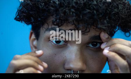 Beschnittene Ansicht eines afroamerikanischen Mannes, der die Augenbraue berührt und isoliert auf Blau auf die Kamera schaut Stockfoto