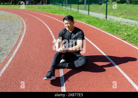 Asiatischer Sportler beim Joggen verletzt, Mann im Trainingsanzug sitzt auf dem Laufband des Stadions und hält Hand an Bein, hat starke Schmerzen in den Muskeln, Bändern, massiert Stockfoto