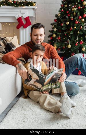 Vater und Sohn sitzen auf dem Boden im Wohnzimmer und lesen Magazin in der Nähe von weihnachtsbaum Stockfoto