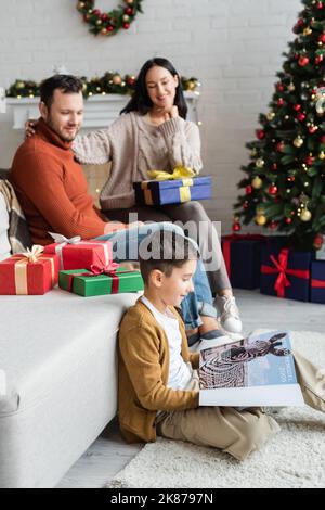 Lächelnder Junge liest auf dem Boden ein Magazin, in der Nähe verschwommener Eltern und Weihnachtsgeschenke auf der Couch im Wohnzimmer Stockfoto