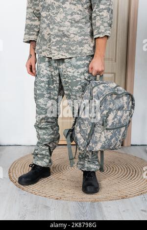 Teilansicht des Militärs in Uniform und Stiefeln, die mit Rucksack in der Nähe der Eingangstür zu Hause stehen Stockfoto