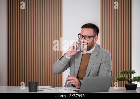 Fröhlicher Unternehmer oder Geschäftsmann, der ein Gespräch am Telefon führt, in seinem Büro sitzt. Stockfoto