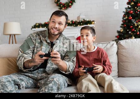 KIEW, UKRAINE - 21. SEPTEMBER 2022: Aufgeregter Junge und Vater im militärischen Uniformspiel mit Joysticks zu Hause Stockfoto