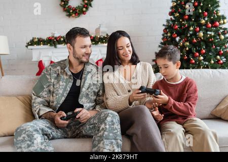 KIEW, UKRAINE - 21. SEPTEMBER 2022: Lächelnder Militärangehöriger schaut auf Frau, die dem Sohn Joystick gibt Stockfoto