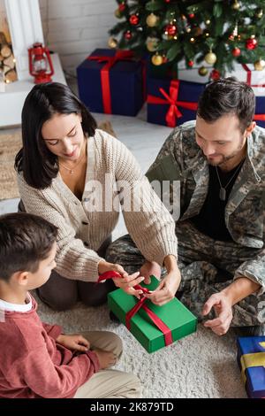 Frau, die zu Hause in Militäruniform ein Band in der Geschenkbox neben Sohn und Ehemann bindet Stockfoto