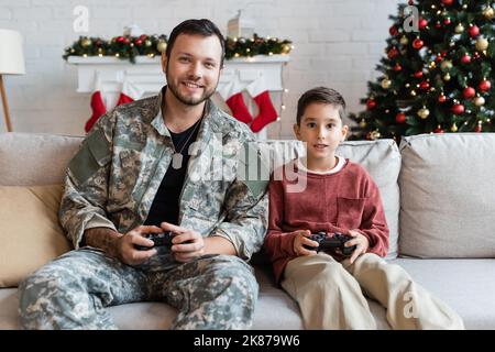 KIEW, UKRAINE - 21. SEPTEMBER 2022: Glücklicher Militärangehöriger spielt Videospiel mit Sohn zu Hause Stockfoto