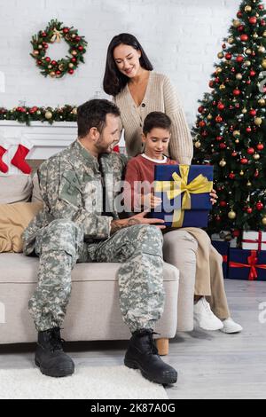 Fröhliche Frau, die den Sohn beim Öffnen der Geschenkbox in der Nähe des Vaters in Camouflage und mit einem geschmückten weihnachtsbaum ansieht Stockfoto