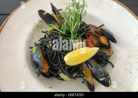 Draufsicht auf schwarze Spaghetti mit saftigen Muscheln, serviert auf Restaurantplatte mit Mikrogemüse und Zitronenkeil Stockfoto