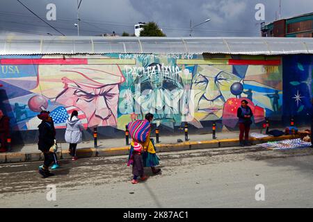 Aymara Dame, die an einem Wandgemälde vorbeiläuft, das Pachamama (Mutter Erde) als traurige alte Frau zeigt, flankiert von Donald Trump und Xi Jinping, La Ceja, El Alto, Bolivien Stockfoto