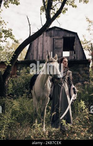 Junge Frau im Bild der alten Reiter Krieger steht in der Nähe von weißem Pferd mit Schwert in der Hand vor dem Hintergrund von Haus und Wald. Stockfoto