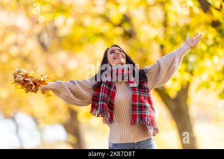 Eine glückliche junge Frau in einem Park wirft während eines indischen Sommers Blätter und genießt den Herbst. Stockfoto