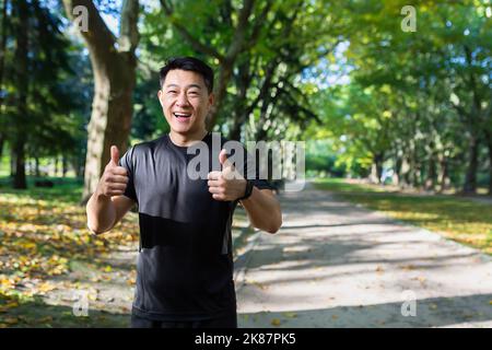 Portier des glücklichen Fitness-Trainers im Park, asiatischer Mann lächelt und schaut auf die Kamera, zeigt die Finger positiv auf, Mann macht Sport im Trainingsanzug an sonnigen Tagen in der Nähe von Bäumen. Stockfoto