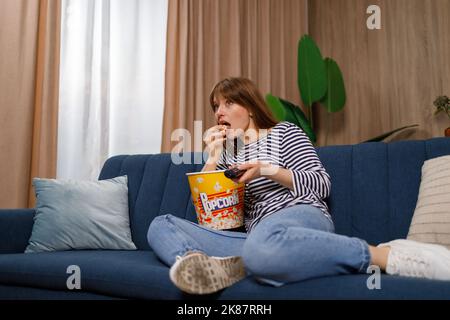 Junge Frau mit Fernbedienung, die Fernsehen schaut und Popcorn auf der Couch isst Stockfoto